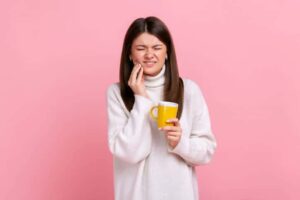 A woman with a dental emergency holding a coffee cup while also holding her face in pain.